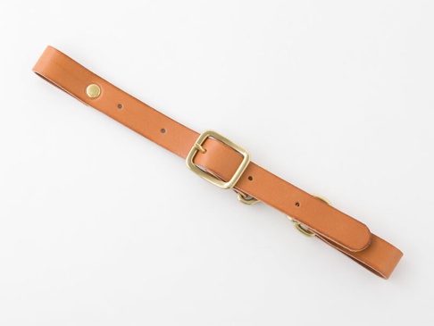 淺棕色皮革製成的背包胸前扣帶，寬度約 2.4 公分，配有黃銅色金屬扣環、壓扣及 D 型環，可用於調整背包肩帶。