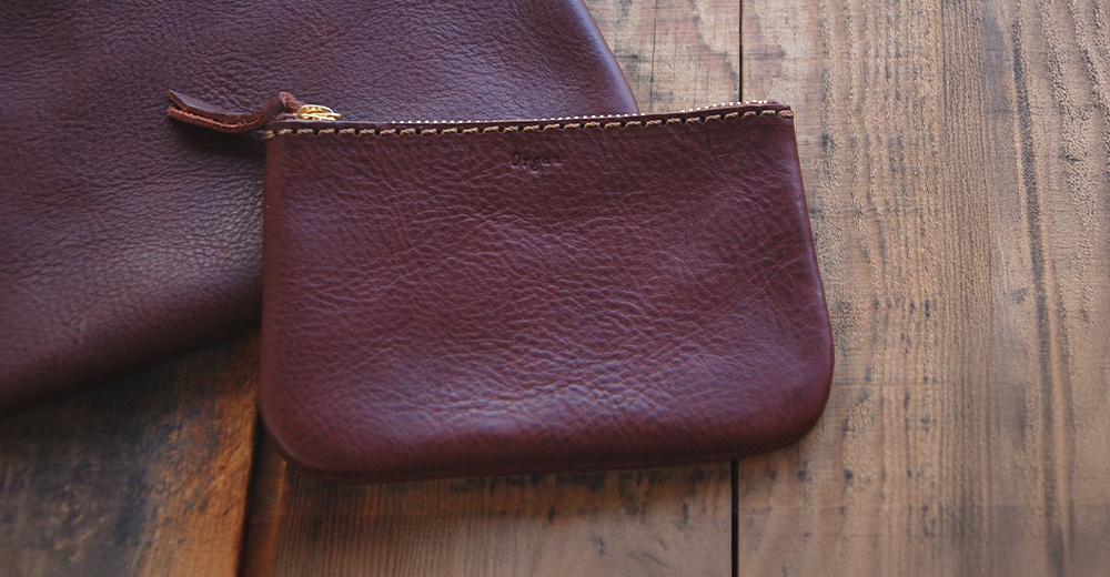 酒紅色職人手作皮革拉鍊式小物收納袋，表面有皮革紋理，搭配金色拉鍊和米色縫線。