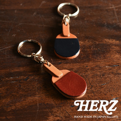 兩個職人手作皮革桌球拍造型鑰匙圈，一個為咖啡色皮革，另一款為咖啡色皮革搭配黑色皮革，均配有金色金屬鑰匙環。