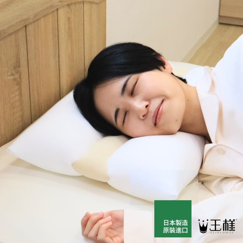 一名女子戴著乳白色睡衣，躺在床上的王樣檜木清香枕上，臉上帶著安詳的微笑，眼睛閉著，似乎睡得很香甜，枕頭的側面露出檜木內芯的米黃色網狀材質，背景是淺木色的床頭板。