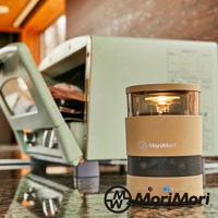 卡其色圓柱形無線藍牙音響，頂部有透明玻璃罩和暖黃色LED燈，中間有網狀喇叭孔，品牌標誌為「MoriMori」。背景為淺綠色復古造型的物品。