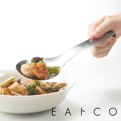 銀色不鏽鋼服務餐匙，匙面盛有鮮蝦、扇貝與花椰菜等料理，由手持，背景為白色，下方有英文字母「E A T C O」標誌。