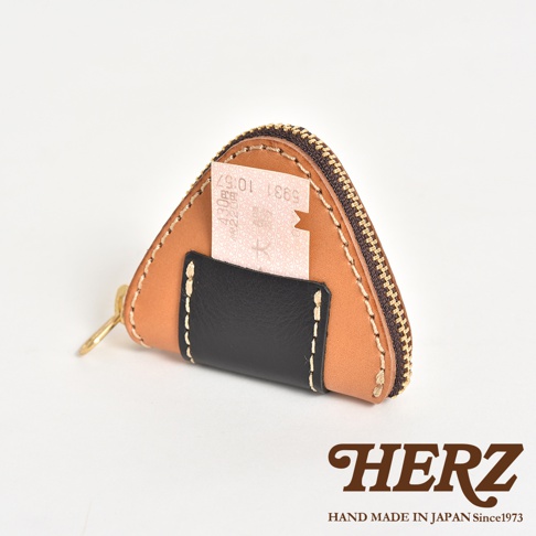 一個日本HERZ職人手作的三角飯糰造型皮革零錢包，以淺棕色皮革製成，正面縫有一個黑色皮革卡槽，卡槽中插著一張車票。錢包邊緣有精緻的粗縫線裝飾，並配有拉鍊開口，右下角印有HERZ品牌名稱及相關標語。