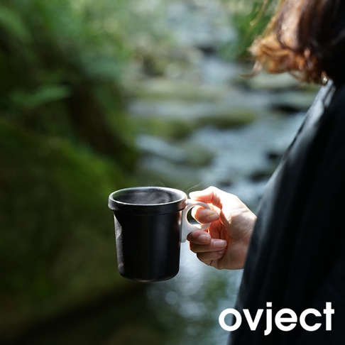 黑色琺瑯馬克杯，帶有白色手把，由人手持著，背景為模糊的綠色自然景觀和流水。