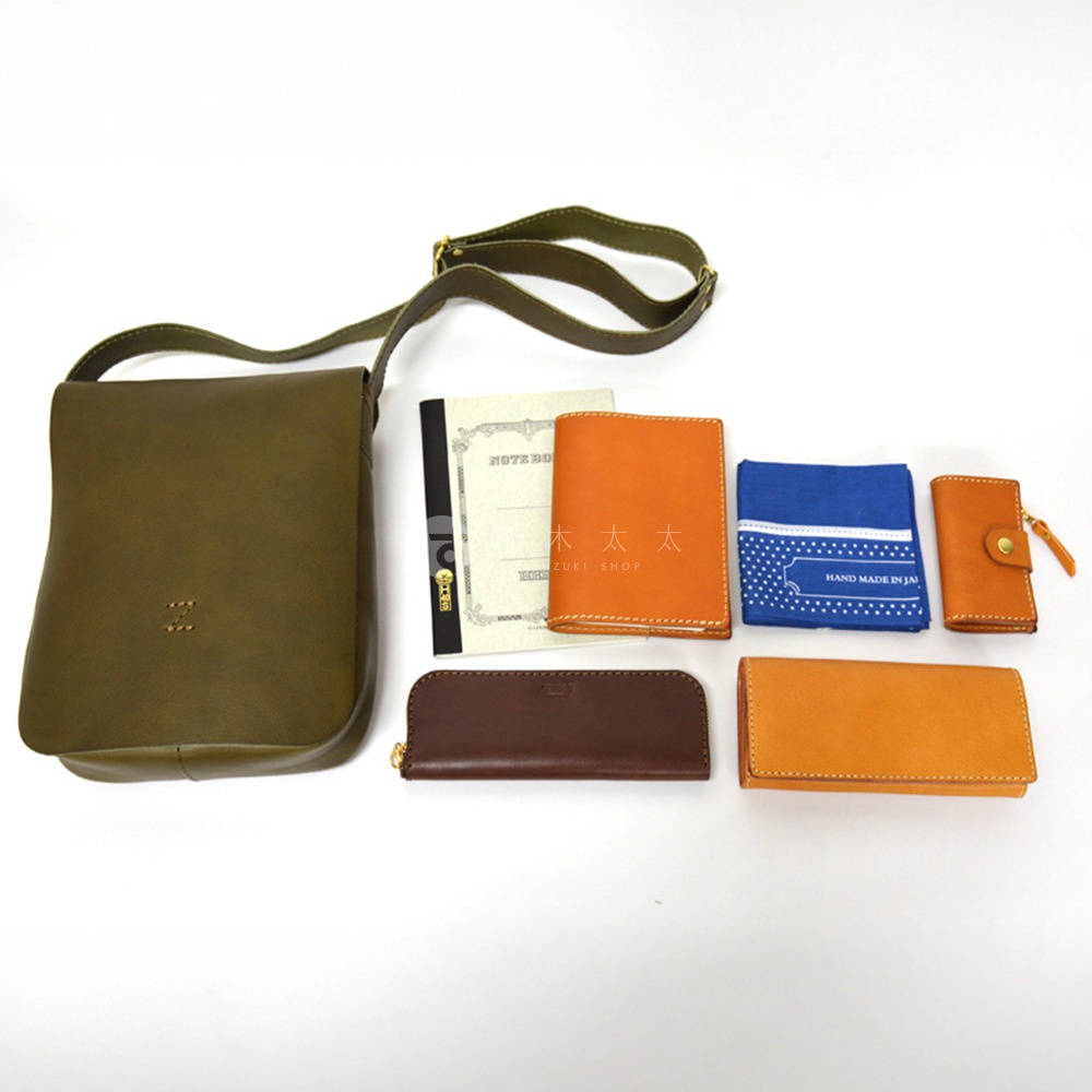 一組日本 HERZ 品牌皮革製品，包含一個橄欖綠色的軟皮斜背包，一個棕色長夾，一個橘色皮夾，以及一本筆記本和一個藍色印花手帕。