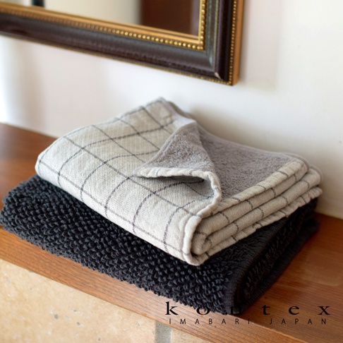 兩條有機棉毛巾，一條為草木染的淺灰色格紋，另一條為深炭灰色，堆疊在木質架上。