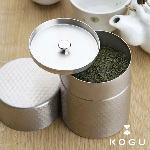 日製18-8不鏽鋼錘目紋茶葉罐，銀色，罐身與蓋子均有手工錘打紋路，罐內裝有綠色茶葉，傾斜的蓋子露出部分茶葉，旁邊另有同款的蓋子。
