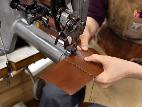 職人正在使用工業縫紉機，縫製一個光滑的棕色皮革三折式迷你短夾。