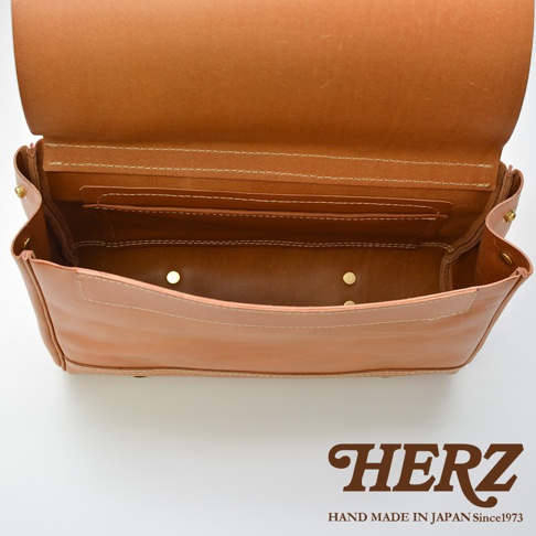 HERZ 品牌標誌，下方為 HAND MADE IN JAPAN Since1973，圖片中央為打開的棕褐色皮革側背包內部，可見其夾層與縫線細節。