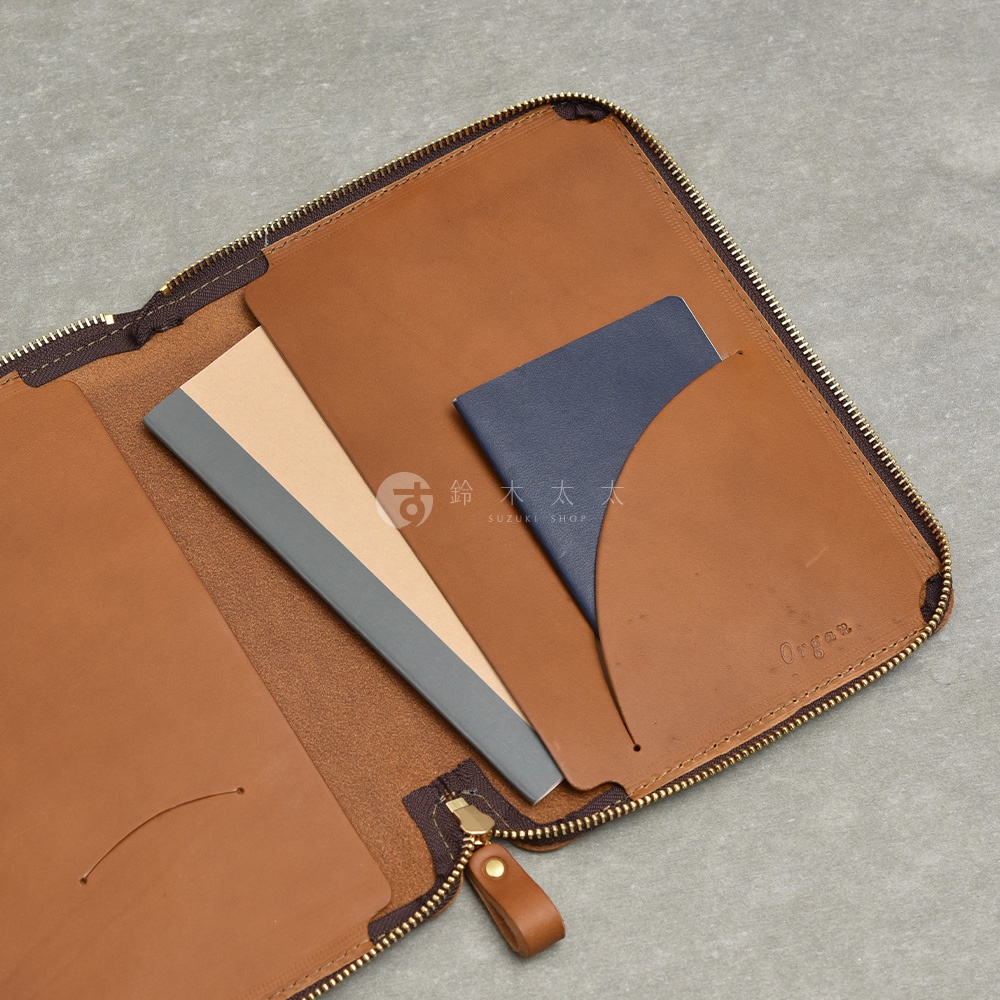 打開的棕色皮革記事本收納包，內部有兩個筆記本和一個藍色護照，右側有一個印有 Organ 字樣的皮夾。