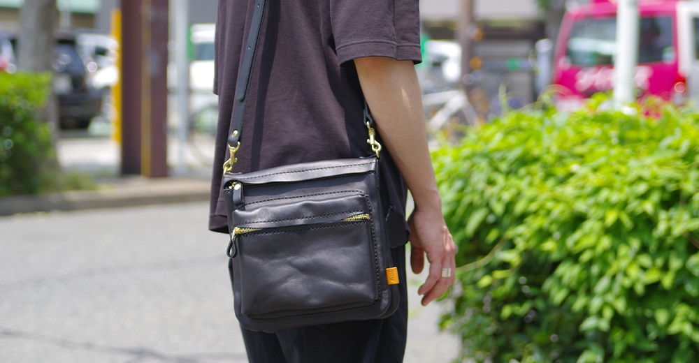 職人手作黑色皮革翻轉直式斜背包，採用光滑皮革材質，設有金色拉鍊和鉤扣，可肩背或斜挎。