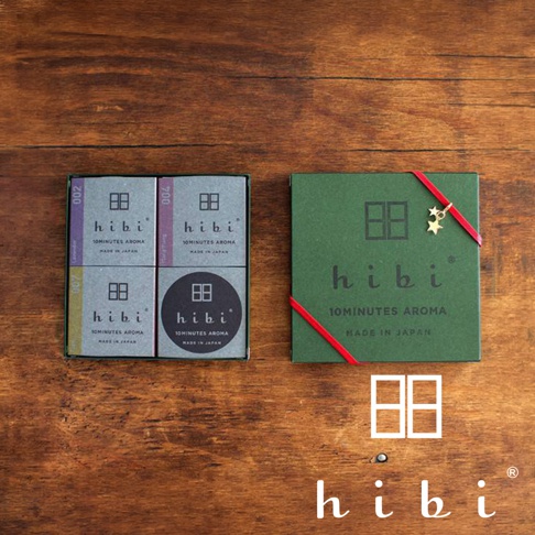 深褐色的木質背景上，擺放著一盒 hibi 聖誕限定版香氛火柴禮盒及其內容物。左側呈現四個標註有 hibi 字樣的香氛小盒，右側為繫著紅色緞帶並綴有星星掛飾的深綠色禮盒外盒，下方則以白色呈現 hibi 的品牌商標及其經典的方形圖示。