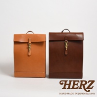 兩款職人手作皮革後背包，一款為淺棕色，另一款為深棕色。兩款皆為紙袋造型，採用皮革材質，配有金屬鉤扣開闔設計。