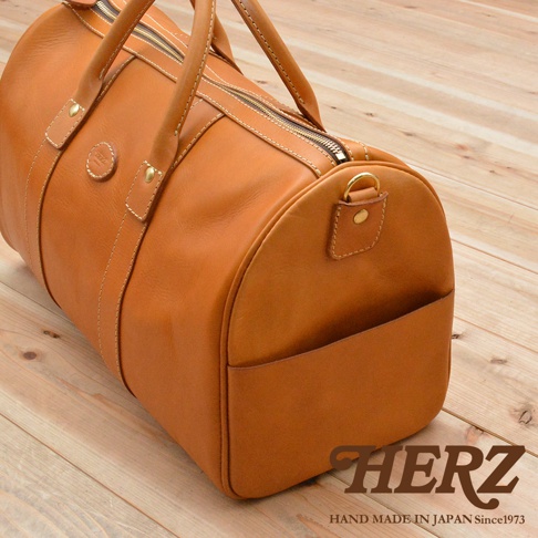 職人手作的棕色皮革波士頓包，具有圓筒形輪廓、頂部拉鍊開口、雙手提把、側邊插袋，以及金屬D型環肩帶連接點，搭配白色縫線。