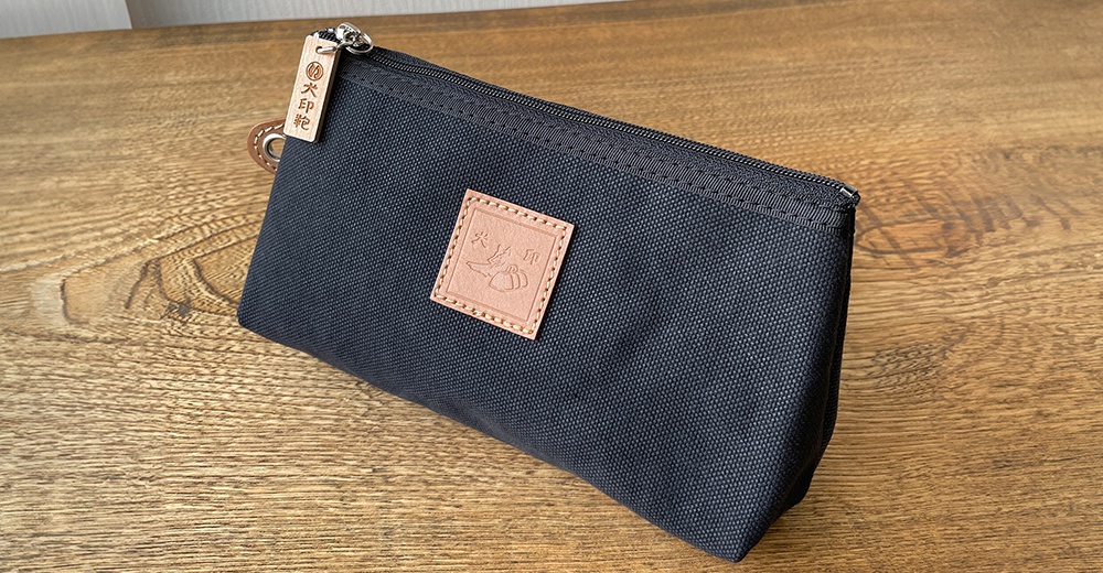 黑色石洗帆布製成的小型收納包，包身有一個方形皮革標籤，上面有燙金圖案，拉鍊頭繫有木製吊牌，吊牌上印有中文。