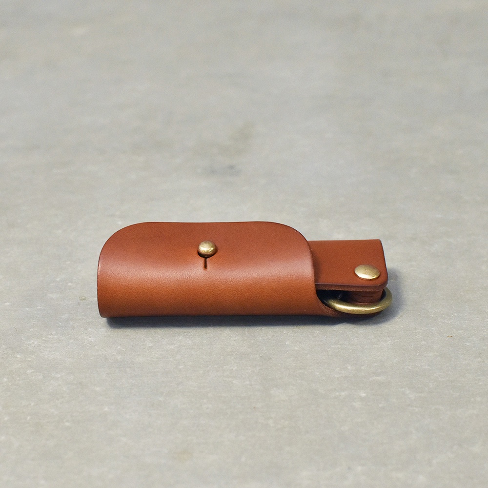 咖啡色皮革製的鑰匙套，擁有圓潤的翻蓋設計，以金色銅釦固定，一側露出鑰匙圈。