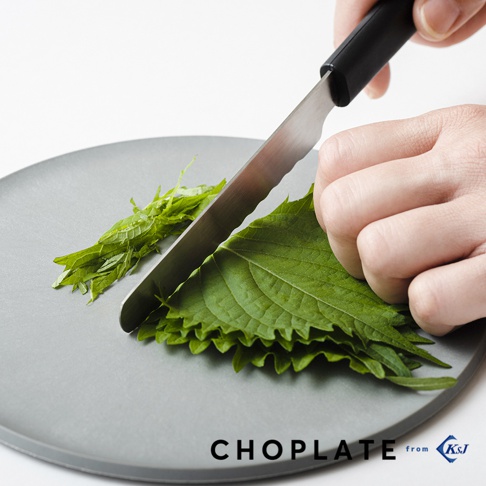 一只手正用一把帶鋸齒刀刃的不鏽鋼刀，在一個圓形灰色砧板盤上切綠色葉菜。