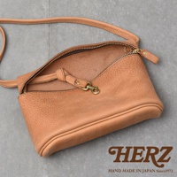 HERZ 品牌標誌，下方為 HAND MADE IN JAPAN Since1973 字樣。 一個淺棕色的皮革斜背包，背包的拉鍊敞開，露出內裡。背包帶和金屬扣環清晰可見。