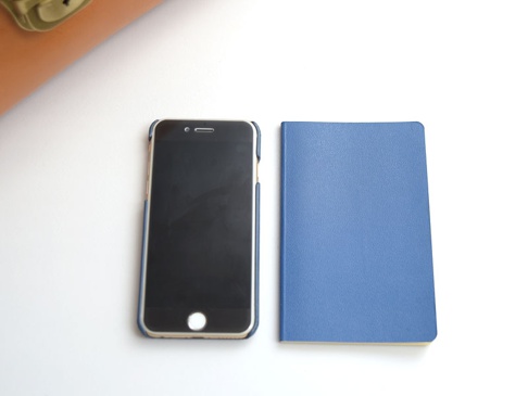 職人手作皮革屋瓦風兩用單肩包 (CW-3)，展示一個深藍色皮革筆記本和一個黑色螢幕的金色手機，手機裝有藍色邊框保護套，兩者並排置於白色背景上。