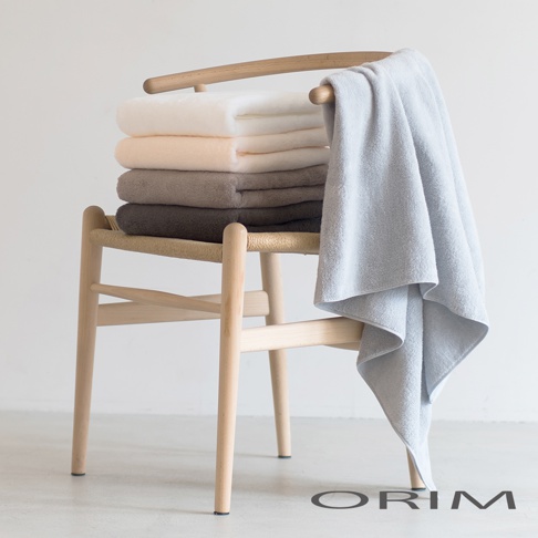 一堆疊放的 PLUME 今治浴巾，顏色由上至下為米白、淺粉、灰褐色、深灰。一條淺藍色浴巾掛在木椅扶手上，材質看起來蓬鬆柔軟。