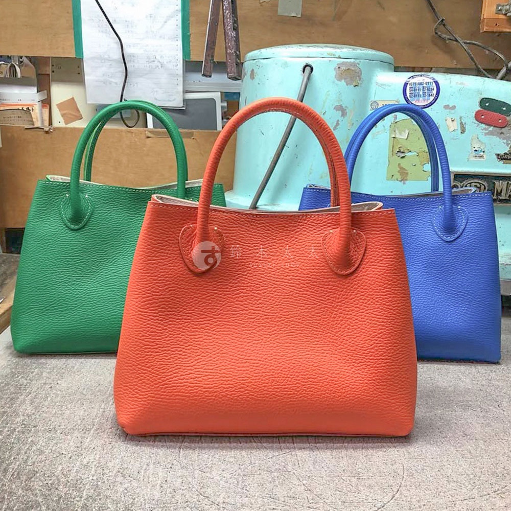 三款京都職人手作手提托特包，分別為亮橙色、草綠色及寶藍色，包身呈現細緻紋理，皮革材質，設計簡約實用。