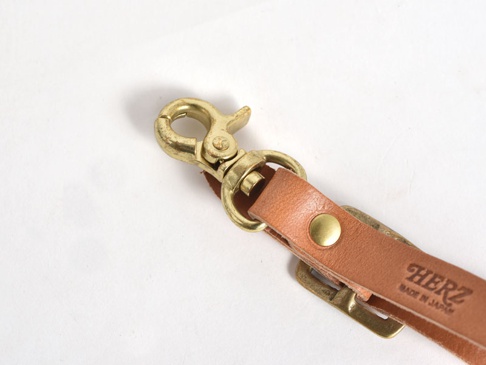 職人手作皮革可調式吊掛帶，採用天然棕色皮革，搭配復古黃銅色金屬扣環和調節器，上面印有「HERZ」品牌標誌及「MADE IN JAPAN」字樣。