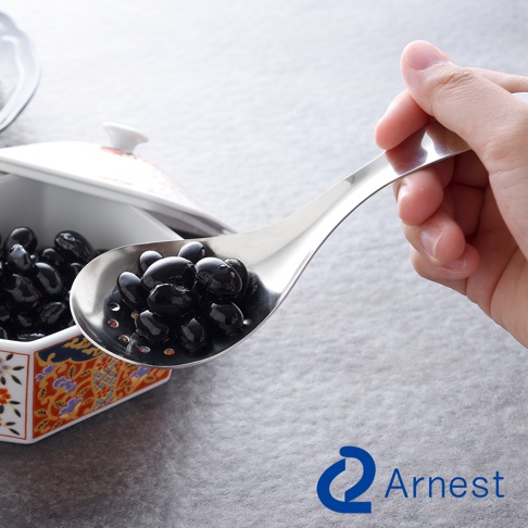 一只手正握著一只不鏽鋼瀝水湯匙，湯匙內裝滿了黑色的豆子，豆子正從湯匙的孔洞中瀝水。湯匙旁是一個裝有黑色豆子的日式風格碗。