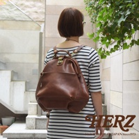 職人手作皮革輕旅行後背包，採用棕色皮革材質，設有束繩開口、金屬鎖扣及前置拉鍊口袋，設計簡約實用。