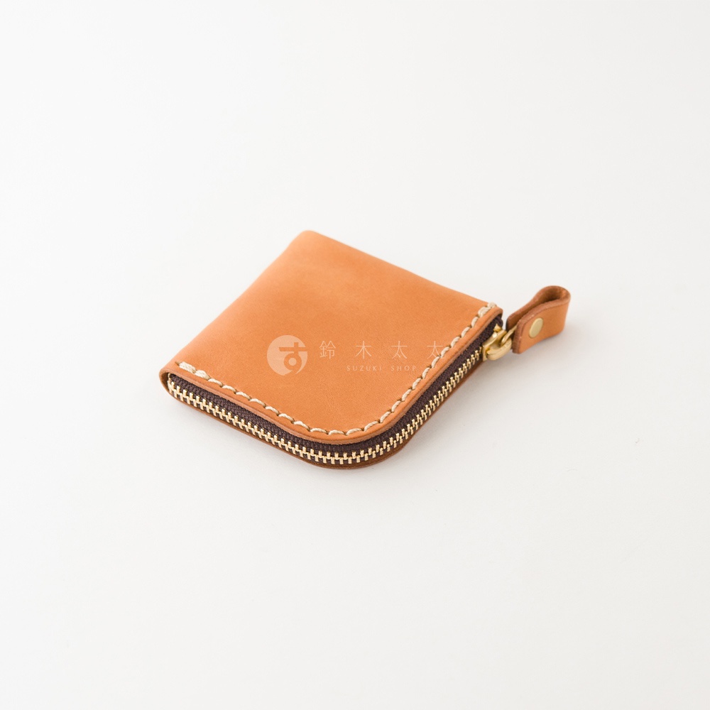 這是一個採用棕色皮革製作的手工零錢包，外觀呈簡約的正方形設計，邊緣處有明顯的手工縫線細節。零錢包配備金屬拉鍊與皮革拉環，表面印有鈴木太太的品牌字樣，整體呈現出精緻的職人手作風格與溫潤質感。