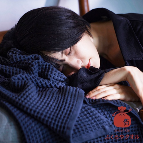 一位女子側臥，臉部埋在深藍色紋理浴巾中，閉著雙眼，表情安詳。