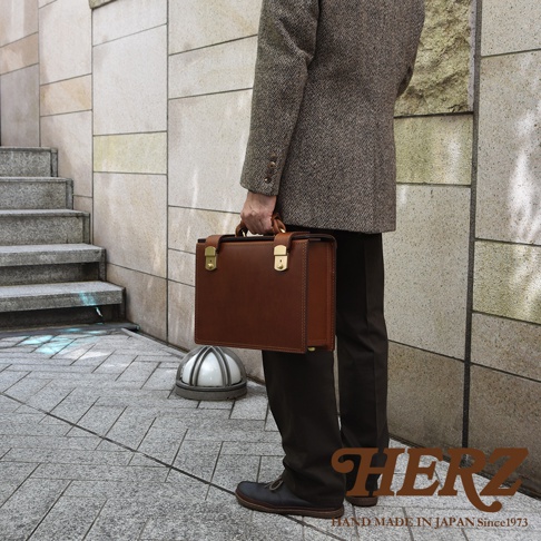 棕色皮革機師箱，配有金色鎖扣和手柄，由一位身穿格紋外套的男士手持，背景為戶外階梯與建築。