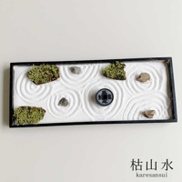 黑框長方形枯山水模型，內有白色細沙，以耙子畫出漣漪紋路，四角和中間各置一塊灰色小石塊，其中央處有一枚有文字的圓形裝飾物，模型兩側各有一簇深綠色苔蘚。