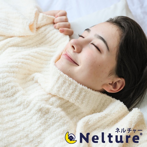 一條奶油色、絨毛材質的連帽毯，帶有羅紋紋理，包裹著一位閉眼微笑的年輕女性。