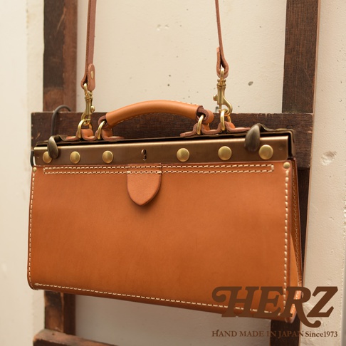 職人手作的復古皮革口金兩用手提包，採用溫暖的棕褐色皮革，搭配古銅色金屬配件及鎖扣，側邊縫線細緻，展現手工質感。