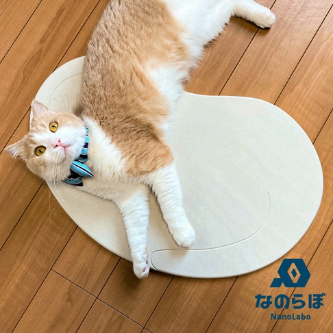 一隻橘白相間、戴著藍色條紋領結的貓咪，趴在淺米色的珪藻土地墊上。地墊呈橢圓形，表面有壓印的貓咪尾巴造型，左下角有「なのらぼ NanoLabo」的標誌。