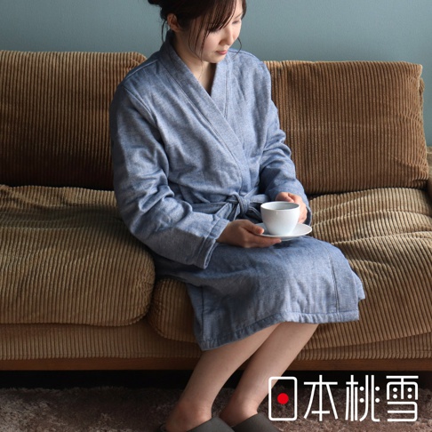 一位女性穿著淺藍色異素材紗質浴袍，坐在棕色燈芯絨沙發上，手持白色茶杯。