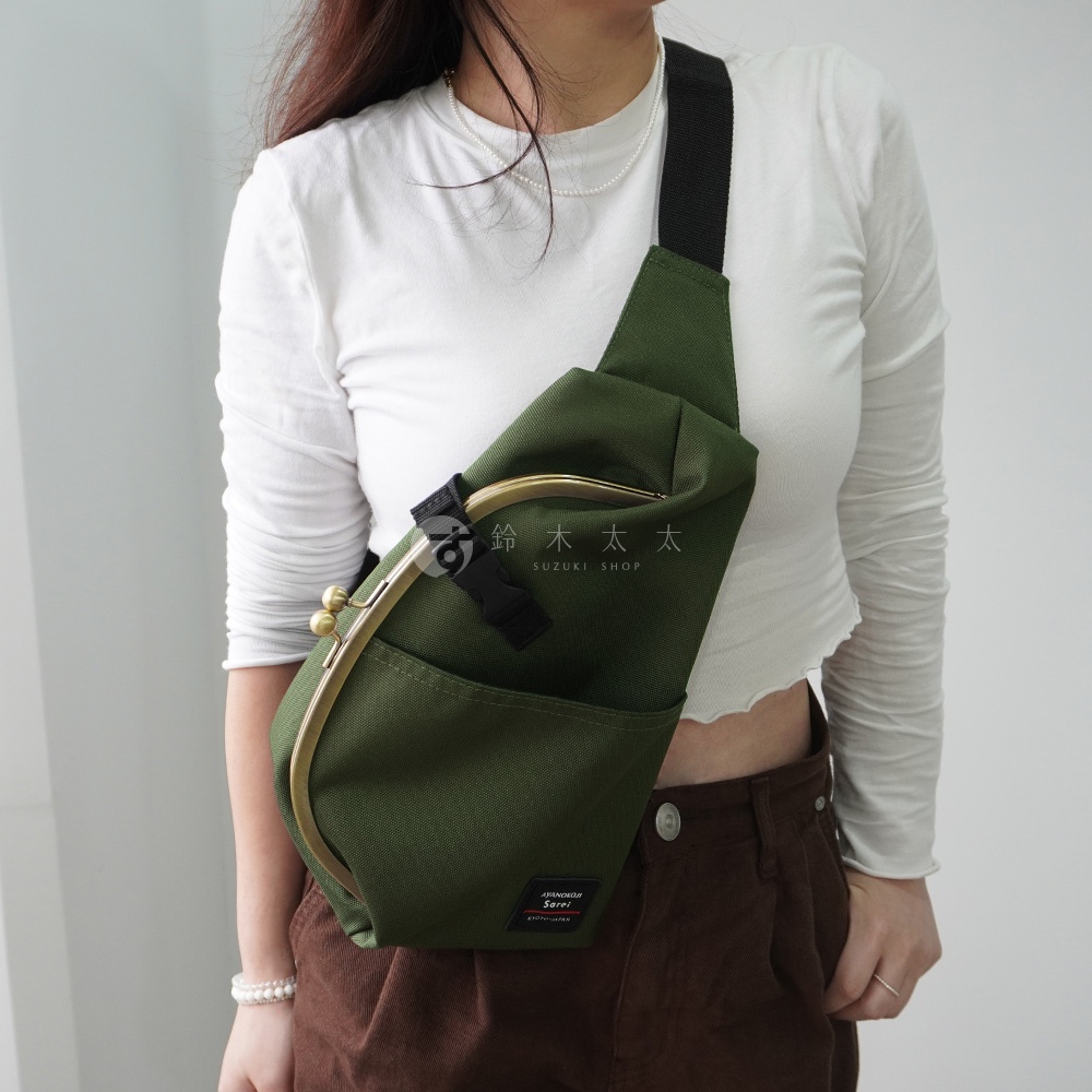 一位身穿白色長袖上衣和棕色燈芯絨褲的女性，斜背著一個綠色的水滴型口金斜背包，背包上有「AYANOKOJI Sarei KYOTO JAPAN」的標籤。