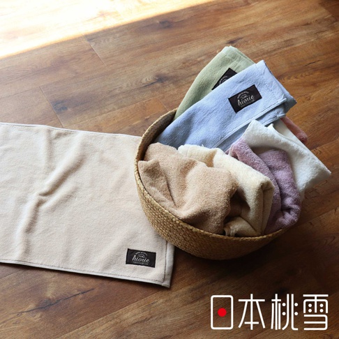 浴墊平鋪在木地板上，顏色為淺米色，表面有細緻的織紋，左下角有黑色標籤。側邊籃子裡有數條不同顏色的毛巾，包括淺綠、淺藍、米白、淡紫和淺棕色，皆有黑色標籤。