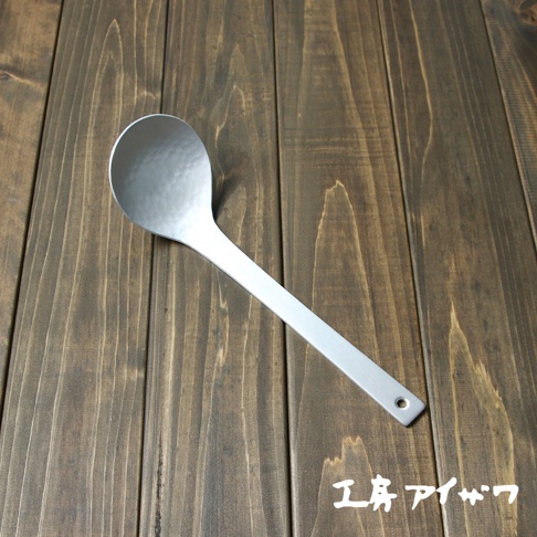 一個銀色鋁製湯勺，勺頭有錘目紋，勺柄末端有一個小圓孔，放置在深棕色木紋背景上。