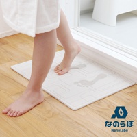 一名女士穿著浴袍，赤腳站在一塊白色的珪藻土地墊上，地墊上有兩個水痕腳印，旁邊是木地板和浴室的入口。圖片右下角有「なのらぼ NanoLabo」的標誌。
