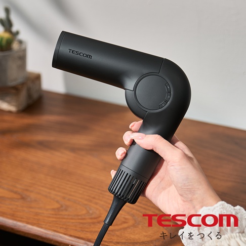 手持黑色 Tescom 摺疊式吹風機，吹風機上有 Tescom 品牌標誌，背景為木質桌面和盆栽植物