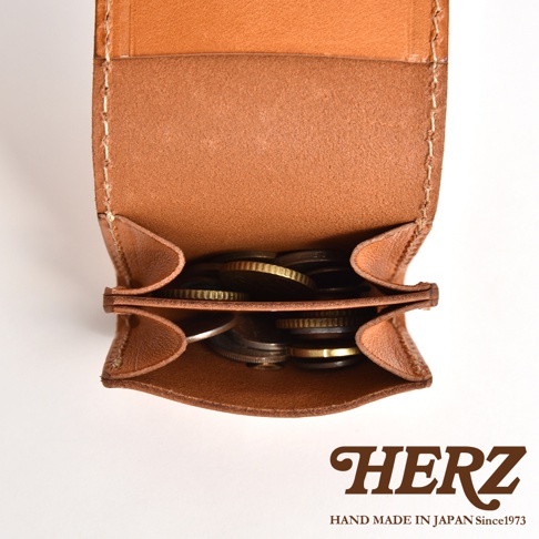 一張由上方俯視拍攝的日本HERZ職人手作皮革零錢包照片，錢包呈現淺棕色調，內部設有分層結構，可以看到硬幣放置其中。圖片右下方標示著HERZ HAND MADE IN JAPAN Since1973字樣。