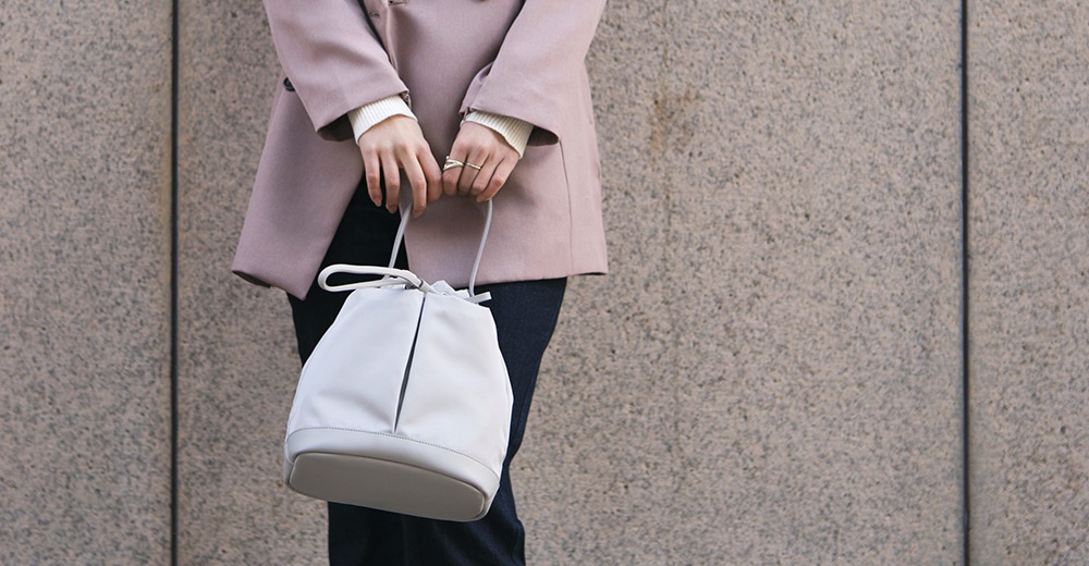 一位女性手持淺灰色的抽繩水桶包，身穿粉色西裝外套和白色針織衫，站在灰色紋理的牆壁前