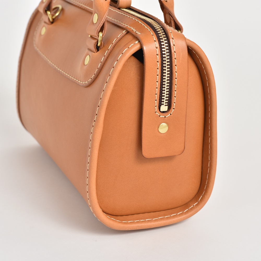 職人手作皮革迷你波士頓包，採用天然皮革材質，呈現溫潤的焦糖棕色。包身線條簡潔，搭配古銅色拉鍊與鉚釘，質感細膩，適合日常攜帶。