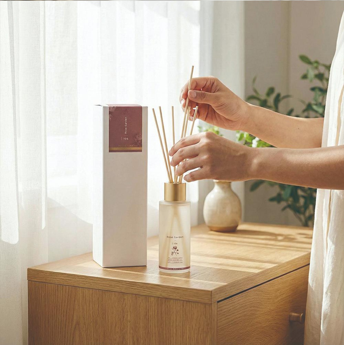 一位女性正在將香氛擴香竹插入裝有「Rose Garden」香味的擴香瓶中，旁邊放著相同品牌的產品紙盒。