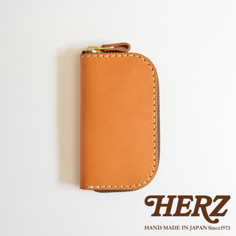 駝色皮革橢圓形拉鍊鑰匙包，配有金色拉鍊及皮革拉環，包身縫線細緻，底部有HERZ品牌標誌，標示HAND MADE IN JAPAN Since1973。