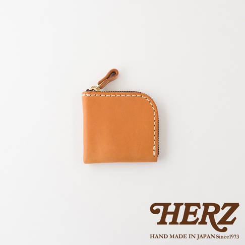 一個淺棕色的日本HERZ職人手作皮革零錢包，採用Stare皮革製成，表面有白色縫線裝飾，配有金屬拉鍊與皮革拉環，右側邊緣呈現圓弧形設計，底部標示著HERZ品牌名稱與手作產地資訊。