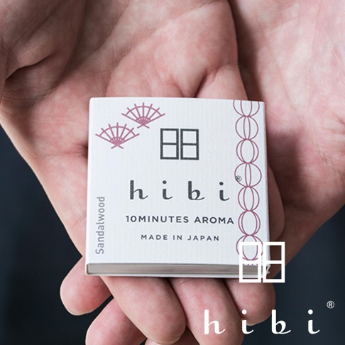 兩隻手心捧著一個白色方形的 hibi 香氛火柴禮盒，包裝上有著日式扇子圖案與簡約的標誌設計，文字標註為 Sandalwood 檀香氣味，整體呈現出精緻的日本傳統工藝質感。