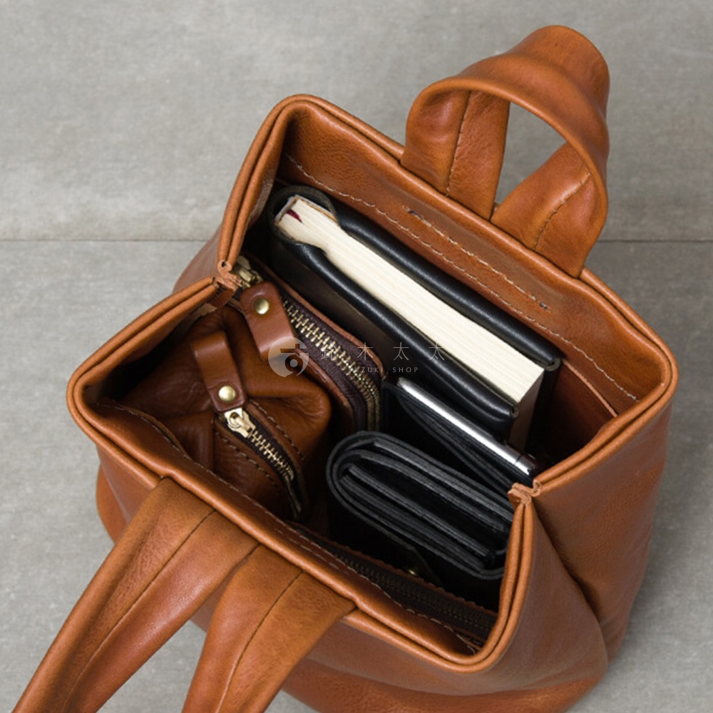 職人手作皮革Organ單柄手提包，呈現溫潤的焦糖棕色調。包內裝有黑色筆記本、錢包和一個小巧的皮革拉鍊零錢包，整體材質為柔軟的牛皮，質感細膩。
