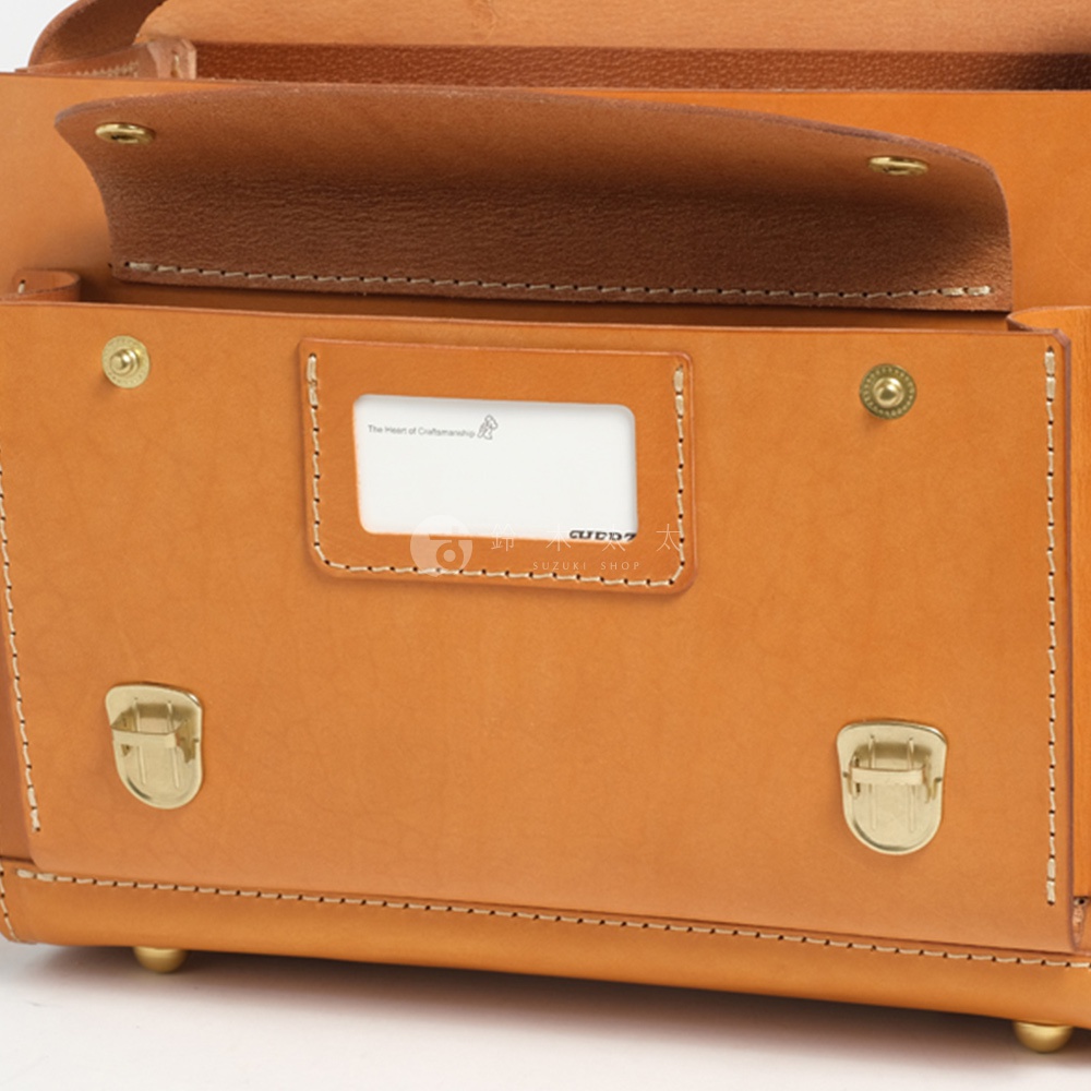 這是一個近距離的特寫，展示了一個淺棕色皮革橫式書包的正面細節。書包由光滑的皮革製成，邊緣有縫線。中間有一個帶有白色卡片的方形皮質標籤，標籤上方印有英文「The Heart of Craftsmanship」和一個小圖案。標籤下方有淺色字體「CUDD7 SUZUKI SHOP」。書包正面有兩個金屬鎖扣，以及底部的四個金屬腳釘。